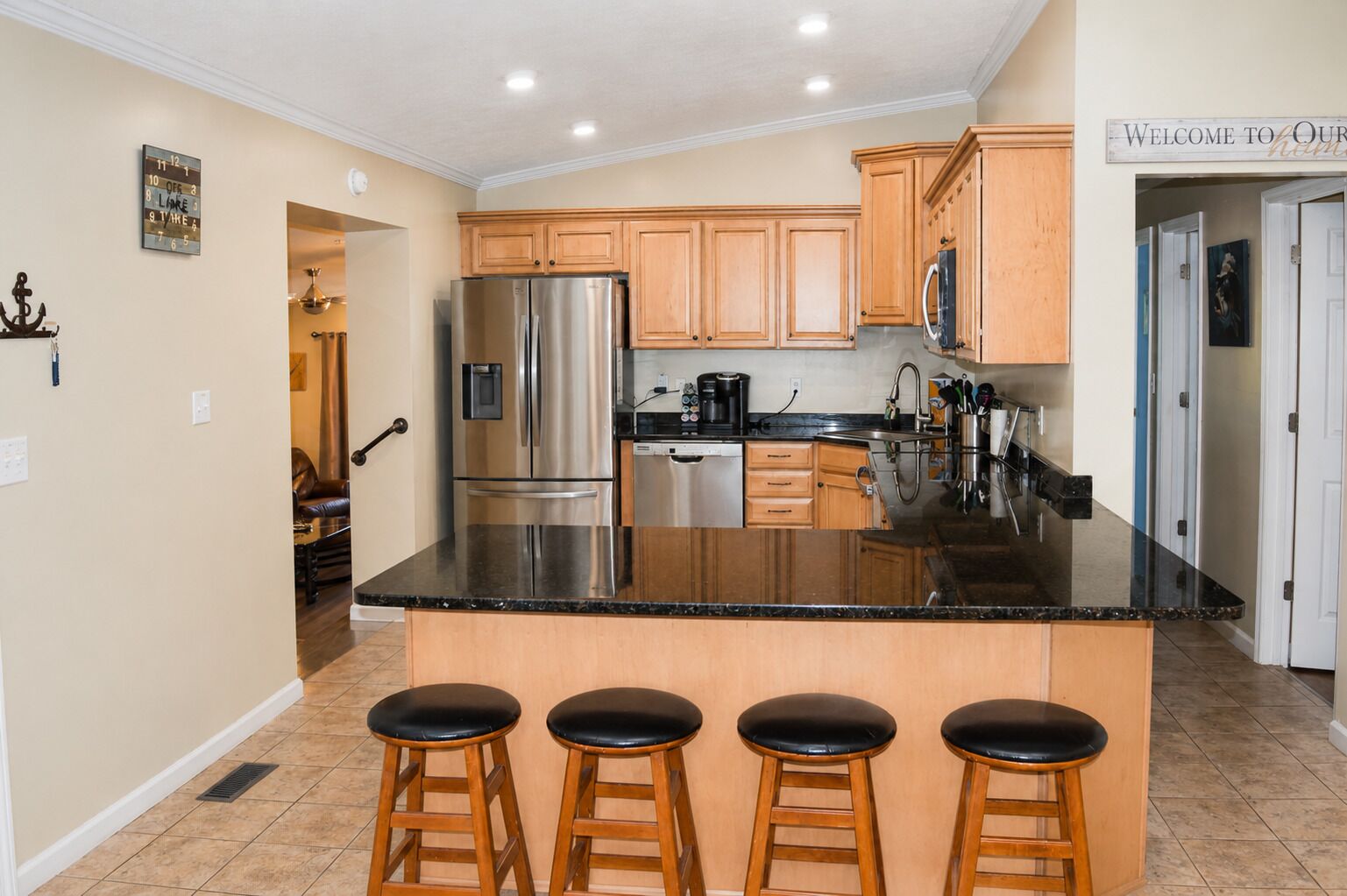 A kitchen with wooden cabinets, a stainless steel refrigerator, a black countertop, and four bar stools.