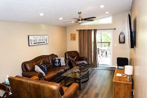 A living room with a leather sofa, a glass coffee table, a ceiling fan, and a sign that reads 'Lake House.' - Keepin' It Reel: Waterfront, 2-Slip Boathouse, EV, Tankless, Park 5 Boats (Guntersville)