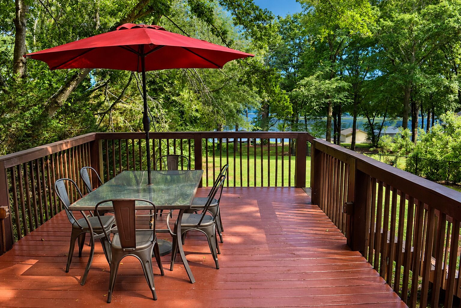 A wooden deck with a glass table and chairs, a red umbrella, and a view of a grassy area with trees.