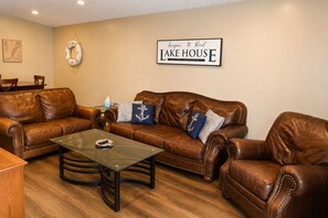 A living room with leather sofas, a glass coffee table, and a sign reading 'Keepin' It Reel LAKE HOUSE ESTABLISHED 2019'. - Keepin' It Reel: Waterfront, 2-Slip Boathouse, EV, Tankless, Park 5 Boats (Guntersville)