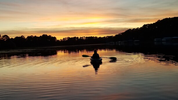 Ein ruhiger See bei Sonnenuntergang, mit der Silhouette einer Person, die ein Boot paddelt.