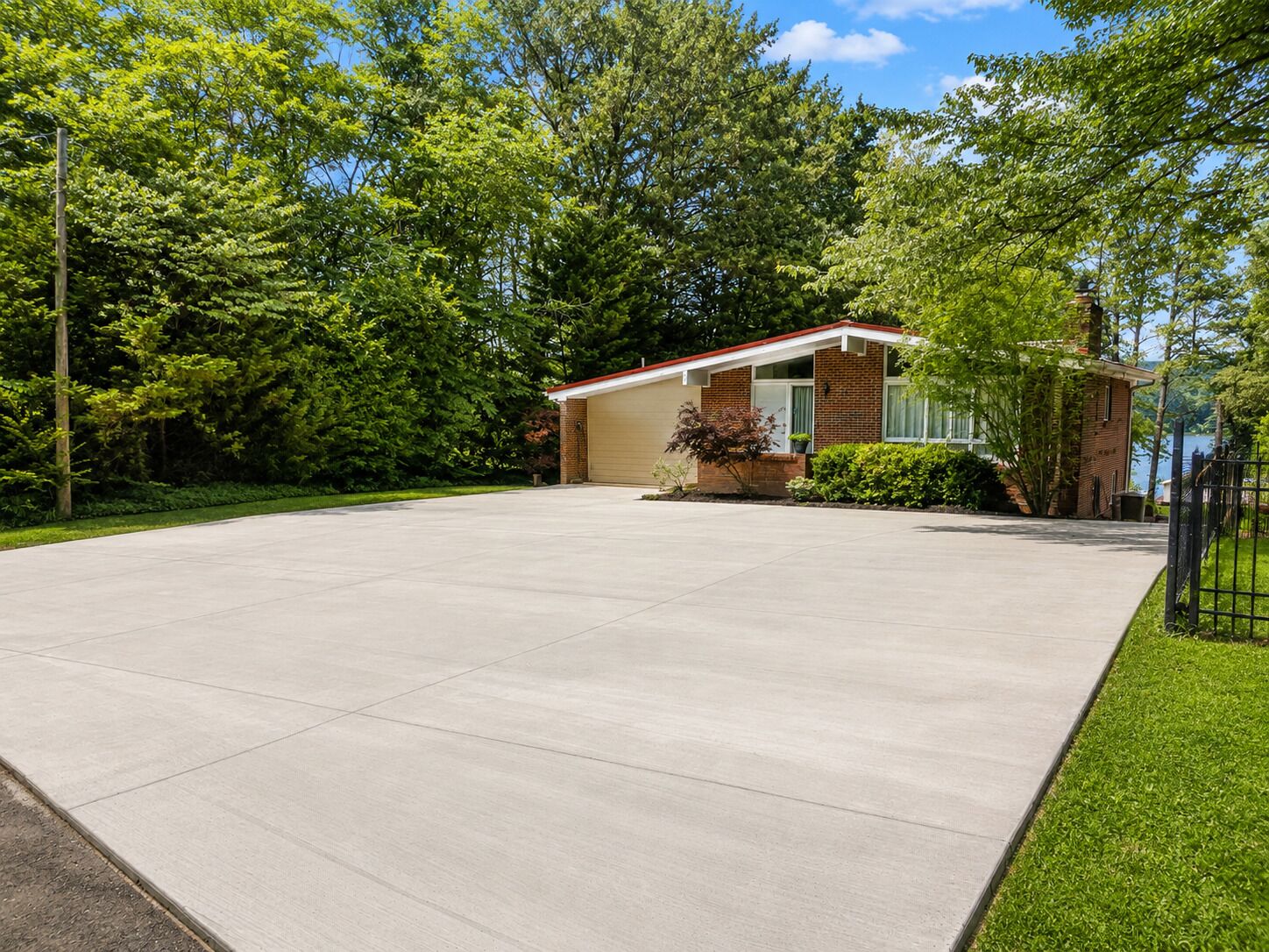 A residential driveway leading to a single-story house with a garage, surrounded by lush greenery and a clear sky.