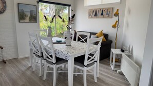 Een eetgedeelte met een tafel voor vier personen, omringd door witte stoelen met zwarte kussens. Er is een raam met uitzicht op groen.