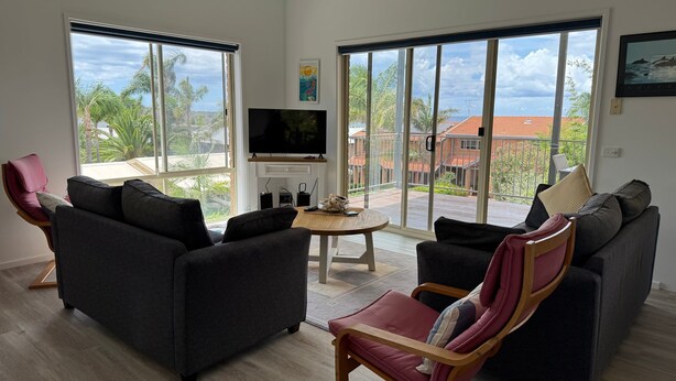 Een moderne woonkamer met twee banken, een salontafel en een televisie. Grote ramen bieden uitzicht op een tropisch landschap.
