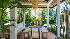 Une terrasse extérieure dotée d’un plafond en bambou, d’une table avec des bancs et de diverses plantes en pot.
