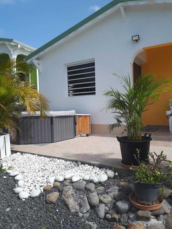 Uma casa com fachada branca, telhado verde e uma pequena janela com barras horizontais. Há plantas em vasos e um caminho de cascalho na frente.