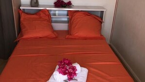 Ein ordentlich bezogenes Bett mit orangefarbener Bettwäsche, einem weißen Kopfkissen und einer Blumenanordnung auf einem weißen Handtuch.