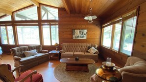 A cozy living room with a wooden ceiling, large windows, a sofa, a red chair, and a coffee table. - GreenGlade Getaway Spacious Cabin & Estate, Lake Winnipeg, Games Room, Veranda (Victoria Beach)