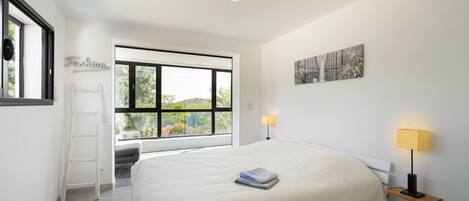 A modern bedroom with a large bed, a ladder, a window with a view, and a wall-mounted lamp.