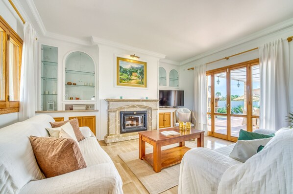 A spacious living room with a fireplace, comfortable seating, and a large window offering a view of the outdoors. - Villa La Romana with private pool in Mallorca (Crestatx)