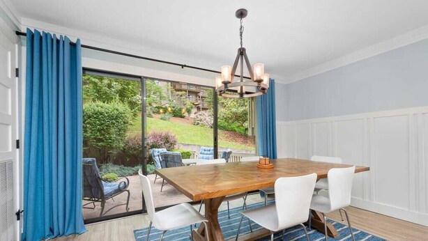 Un comedor con una mesa de madera y sillas blancas, una araña y vistas a un jardín a través de amplios ventanales.