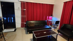 Een woonkamer met een bank, een salontafel en een televisie. Er is een rood gordijn en een witte deur.