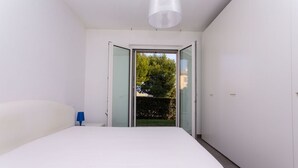 A bedroom with a bed, a nightstand, a blue lamp, and a sliding glass door leading to a garden. - S018 - Sirolo, brand new three-room apartment in stone with right garden (Sirolo)
