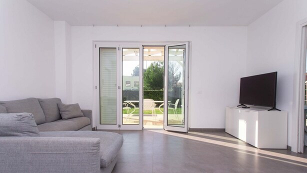 A modern living room with a grey sofa, a flat-screen TV on a white cabinet, and a sliding glass door leading to an outdoor patio. - S018 - Sirolo, brand new three-room apartment in stone with right garden (Sirolo)
