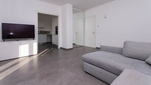 A modern living room with a grey sofa, a flat-screen TV on a white cabinet, and a kitchen area in the background. - S018 - Sirolo, brand new three-room apartment in stone with right garden (Sirolo)