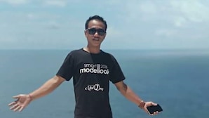 Une personne se tient debout sur un promontoire rocheux, les bras tendus, vêtue d’un t-shirt noir avec une inscription et tenant un smartphone.