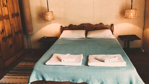 Una cama con una colcha verde, dos almohadas blancas y dos toallas dobladas. Se observa un cabecero de madera, un armario de madera y dos lámparas de mimbre sobre mesitas de noche.