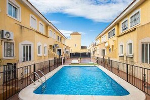 A swimming pool surrounded by yellow buildings with balconies, a clear sky, and a distant tower. - CH Palacios Formentera del Segura (Los Palacios)