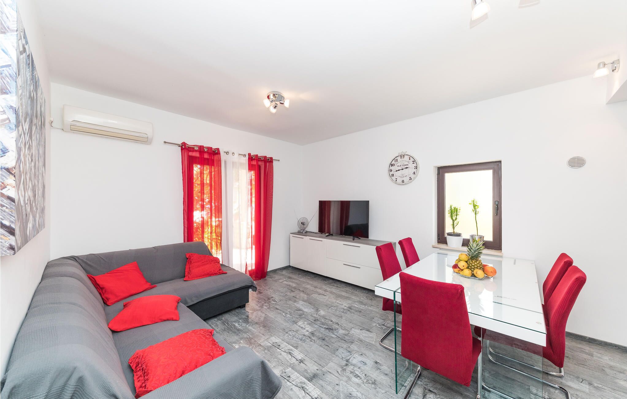 Un moderno salón con un sofá gris, cojines rojos, una mesa de comedor blanca con sillas rojas, un televisor y un reloj en la pared.