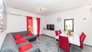 Un moderno salón con un sofá gris, cojines rojos, una mesa de comedor blanca con sillas rojas, un televisor y un reloj en la pared.