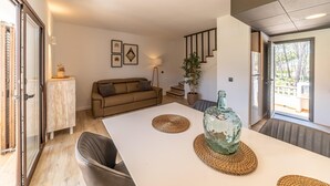 A modern living room with a white dining table, a brown sofa, and a large window. - Gardernia: House for holiday rental with community garden and pool (L'Escala)