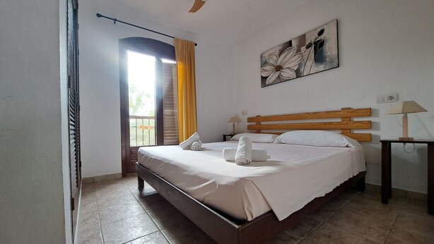 A bedroom with a wooden bed, a ceiling fan, a window with curtains, a painting on the wall, and a nightstand with a lamp. - Es Pujol: Apartment/ flat - Es Pujol (Es Pujol)