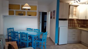 A compact kitchen with a refrigerator, a dining table with chairs, and a wall-mounted shelf with decorative items. - Es Pujol: Apartment/ flat - Es Pujol (Es Pujol)