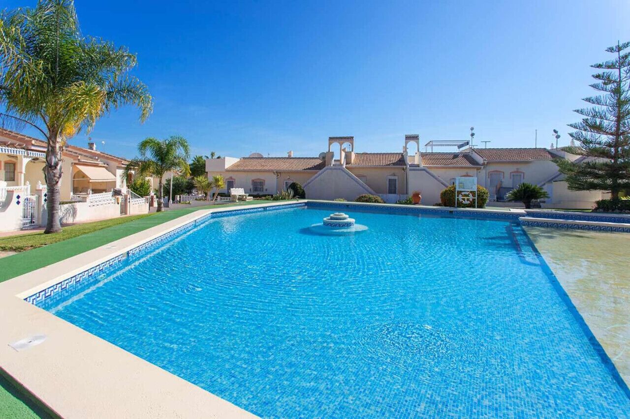 A swimming pool with a fountain, surrounded by palm trees and a building with a tiled roof.