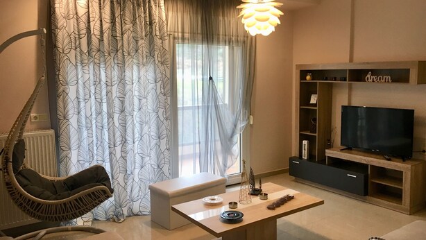 Ein modernes Wohnzimmer mit einer Couch, einem Couchtisch, einem Fernseher und einem großen Fenster mit Vorhängen.