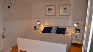 Quarto com uma cama branca, duas mesas de cabeceira com abajures, um radiador e duas molduras na parede.
