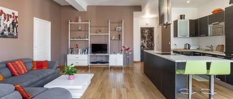 A modern living room with a grey sofa, a white coffee table, and a TV. There is a kitchen area with a bar and stools.