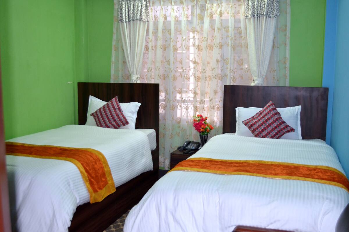 A hotel room with two single beds, each with a wooden headboard, white bedding, and orange throw blankets. There is a small table with a telephone and a vase with flowers between the beds.