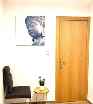 A room with a wooden door, a framed artwork on the wall, a black chair, and a small round table with a plant.