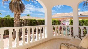 A covered outdoor seating area with a white balustrade, beige cushioned chairs, and a view of a residential area with a palm tree and greenery. - CH Isla Menorca Ciudad Quesada (Ciudad Quesada)