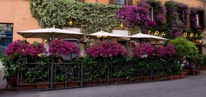 A café with outdoor seating, covered by umbrellas, and surrounded by lush greenery and vibrant flowers.