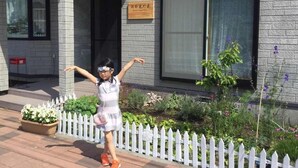 Una bambina in vestito che sta davanti a un edificio con un recinto di legno bianco e un cartello.