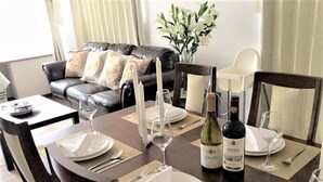 Un tavolo da pranzo apparecchiato con piatti, posate, bicchieri da vino e una bottiglia di vino, con un divano e cuscini decorativi sullo sfondo.