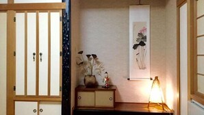Un interno tradizionale giapponese con porte scorrevoli, un piccolo armadietto con un vaso e una figurina, e un'opera d'arte appesa al muro.