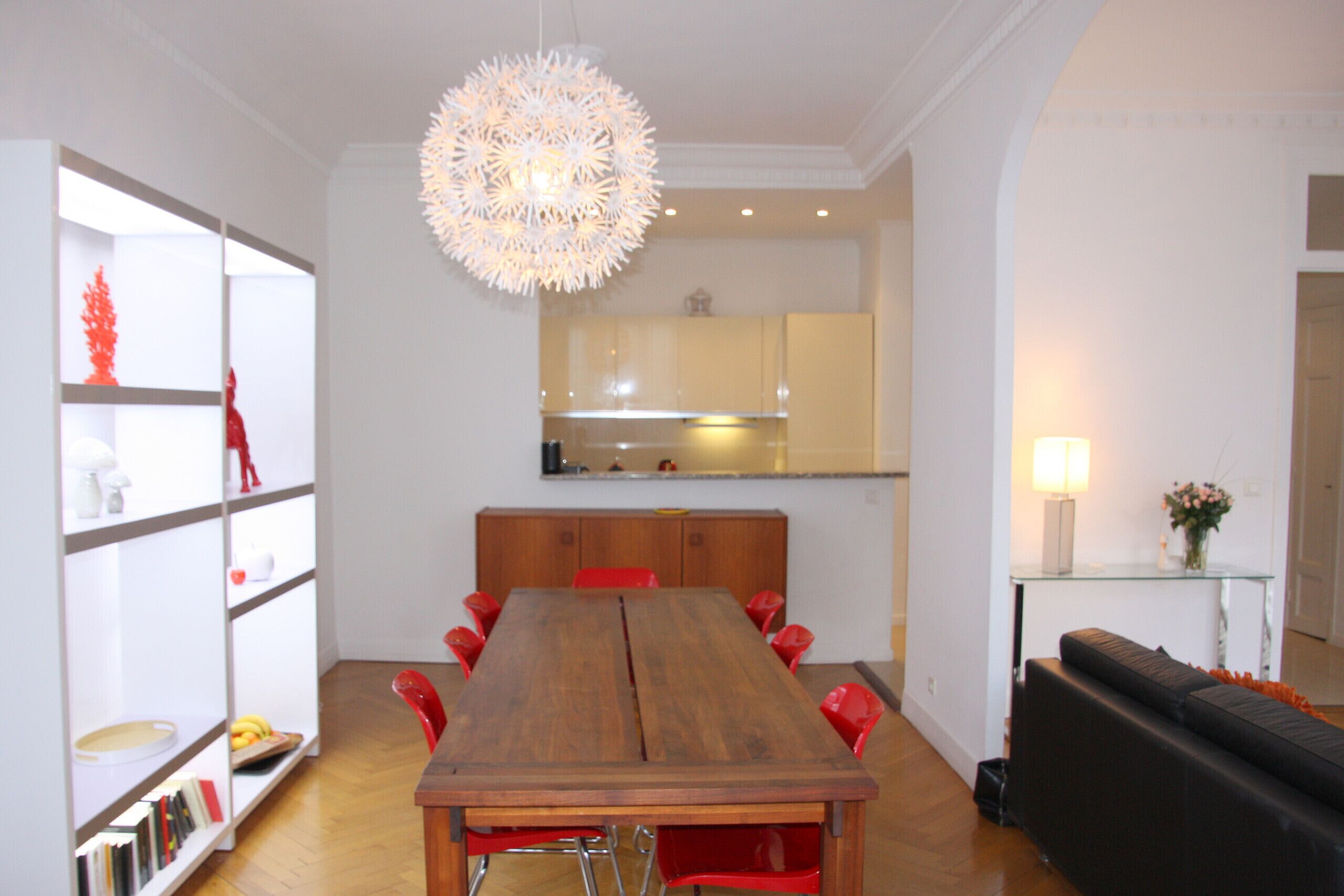 A dining area with a wooden table and red chairs, a black sofa, and a decorative light fixture.
