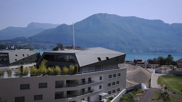 A modern building with a flat roof and multiple balconies, surrounded by greenery and a mountainous backdrop.