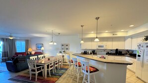 A modern kitchen with white cabinets, a central island with bar stools, and a dining area with a wooden table and chairs. - Chincoteague Getaway on the Bay - Water Front,Pool ,IN TOWN ,LINENS & TOWELS (Chincoteague)
