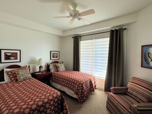 Een hotelkamer met twee bedden, een plafondventilator, een raam met gordijnen, een bank en twee ingelijste foto's aan de muur.