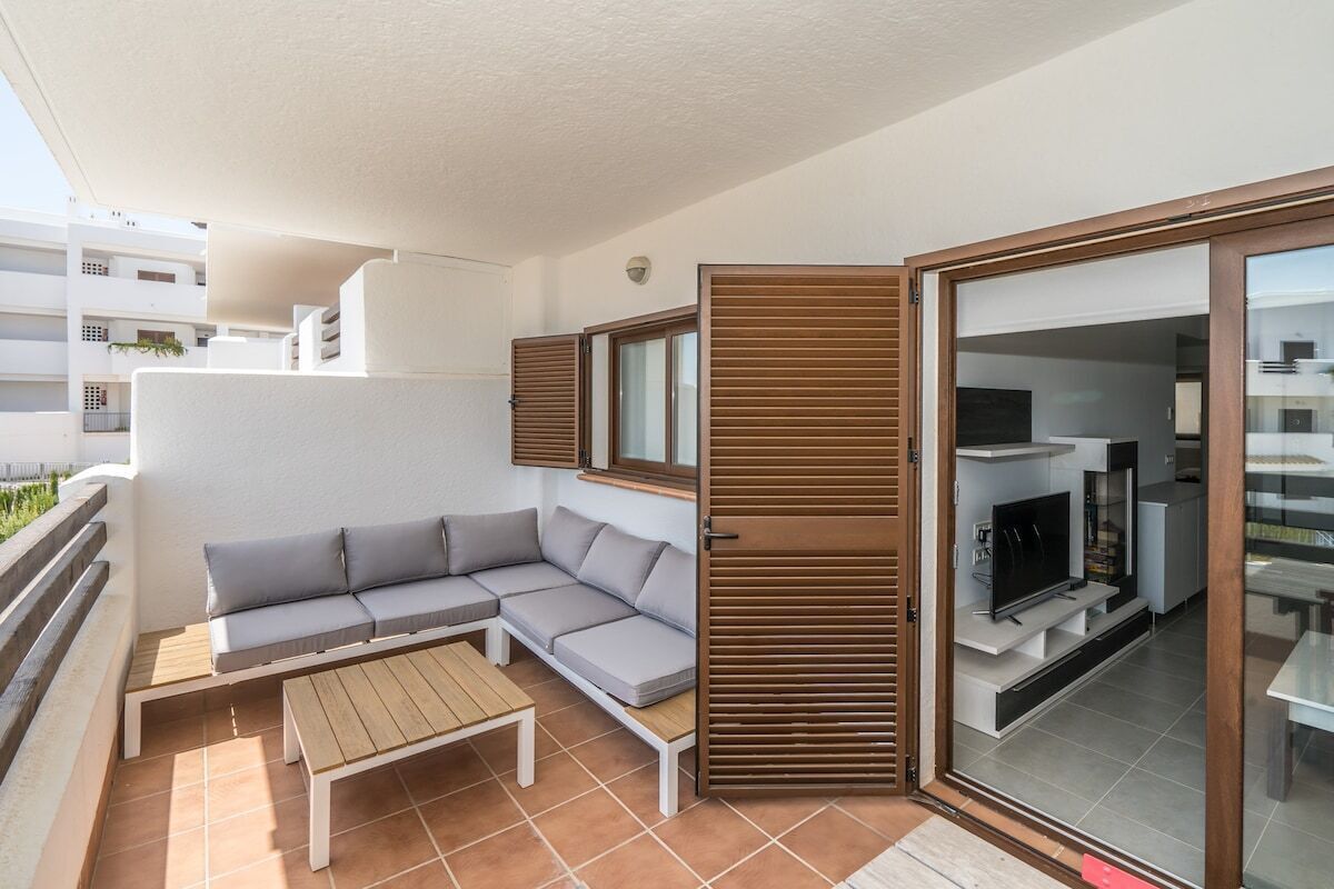 Ein moderner Balkon mit einer grauen Eckcouch, einem Holztisch und einer Glastür, die zu einem Küchenbereich mit einem Fernseher führt.