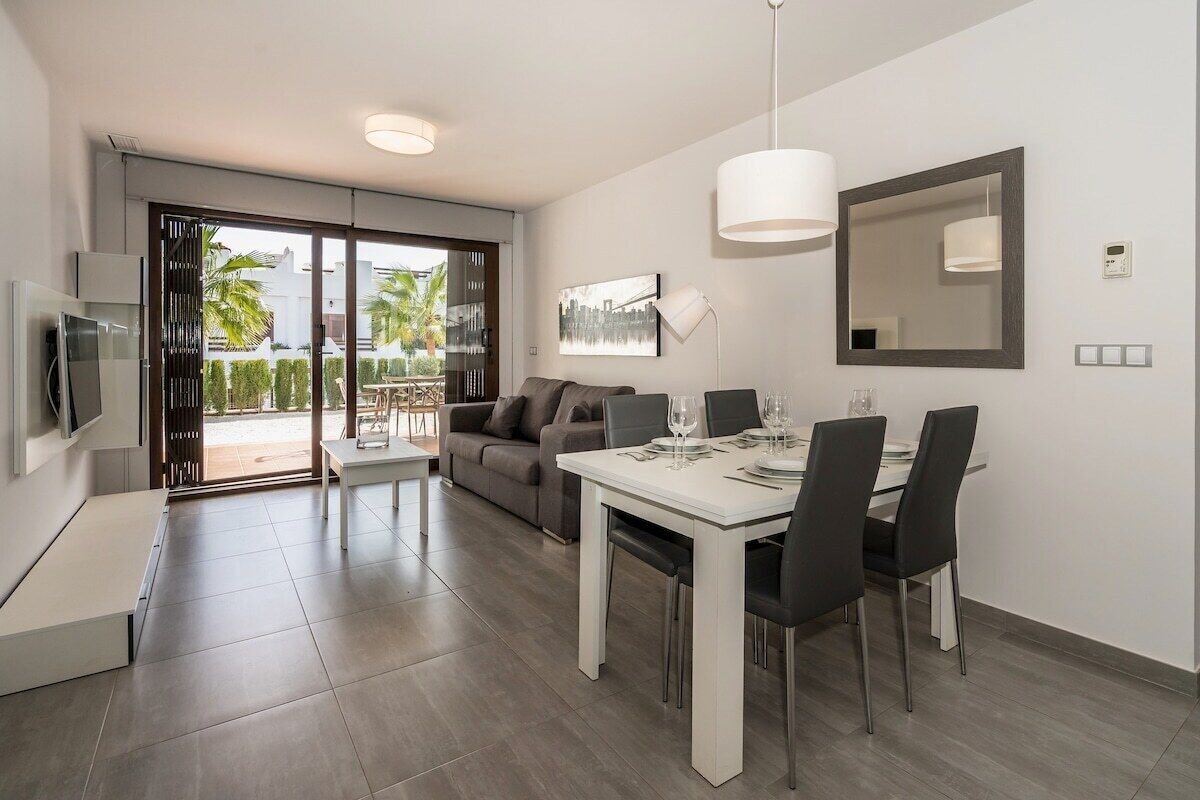 A modern living room with a dining area, a sofa, a TV, and a balcony with a view of palm trees.