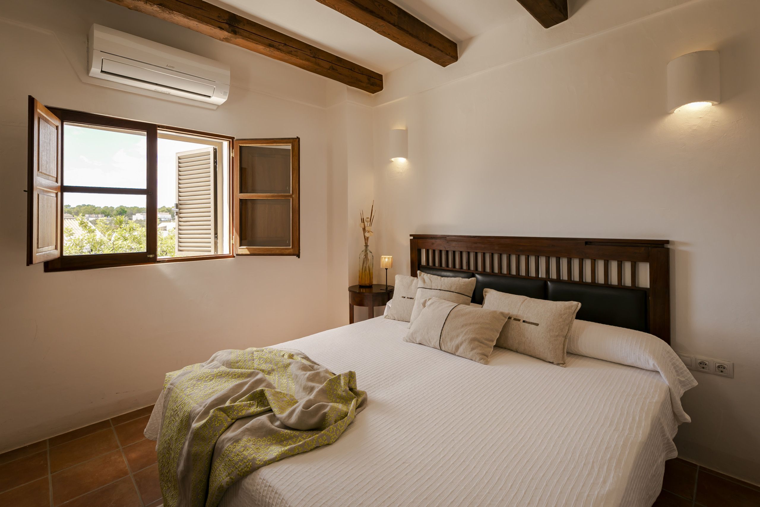 A bedroom with a large bed, a window with shutters, a nightstand, and a wall-mounted lamp.