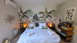 A bedroom with a large bed, two bedside tables with lamps, a wall-mounted air conditioner, a decorative plant, and a framed picture of birds. - Happyfew La petite Acacia (Coti-Chiavari)