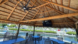 A covered outdoor seating area with a thatched roof, wooden beams, and a ceiling fan. Tables and chairs are arranged for dining or relaxation.