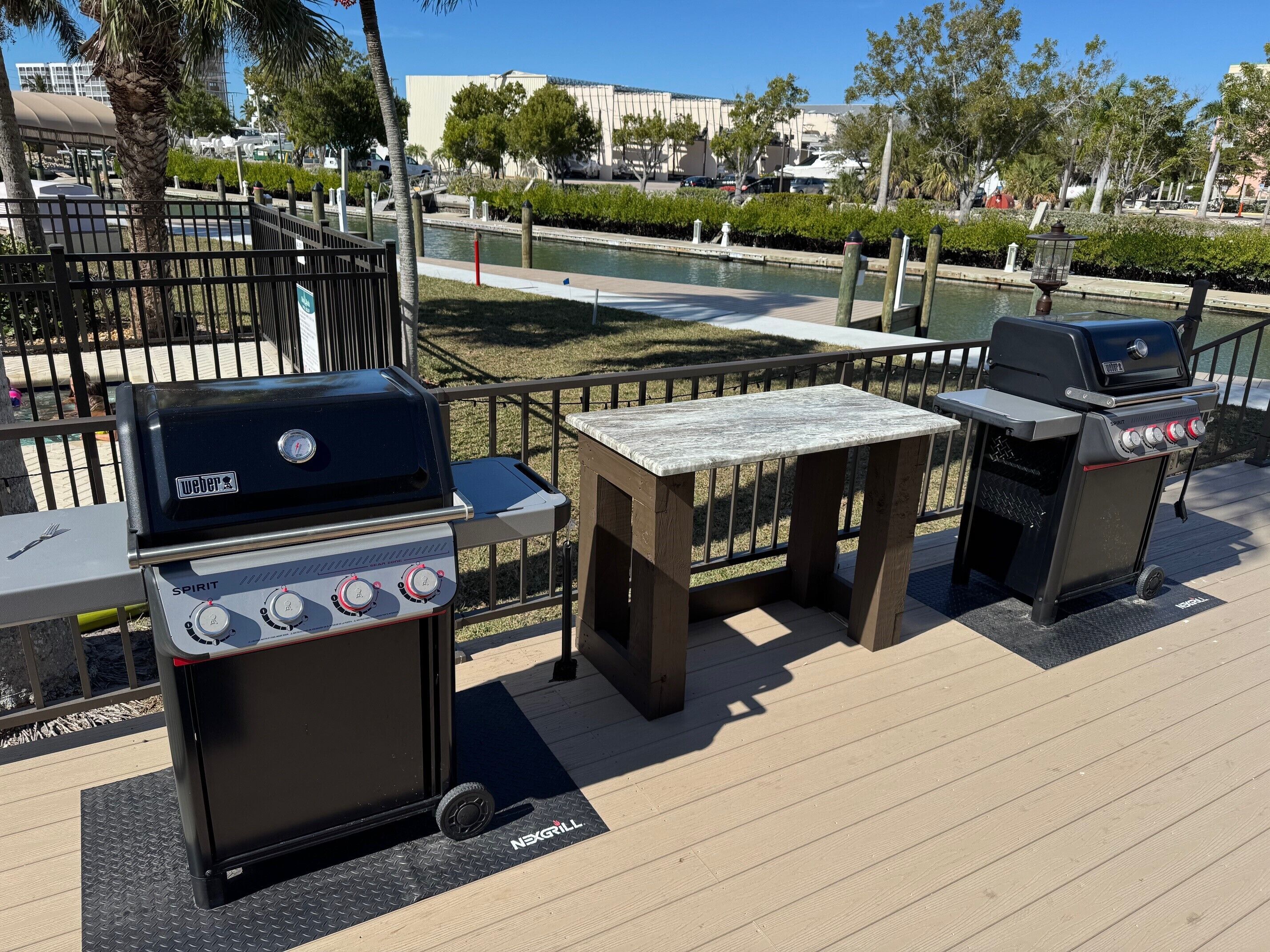 An outdoor patio with two Weber Spirit grills and a wooden table, surrounded by a metal fence and a water feature.