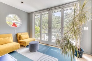 Een moderne woonkamer met een gele bank, een grijze poef, een potplant en een groot raam met uitzicht op bomen en een balkon.
