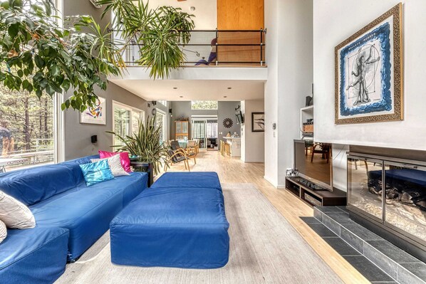 Een ruime woonkamer met een blauwe modulaire bank, een open haard en een groot raam met uitzicht op een balkon.
